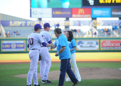 Baseball player in a purple and white uniform shaking hands with a woman on the field, with spectators and digital displays in the background.