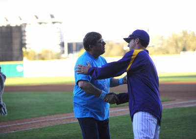 Two men shaking hands on a baseball field, one in a blue shirt and the other in a purple and gold baseball uniform.