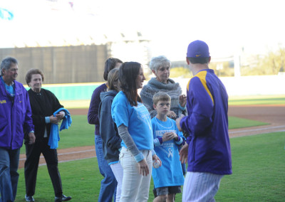 A baseball player in a purple jersey interacts with a group of fans wearing light blue shirts on a baseball field.