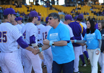 A man in a blue t-shirt shakes hands with baseball players in purple uniforms on a field.