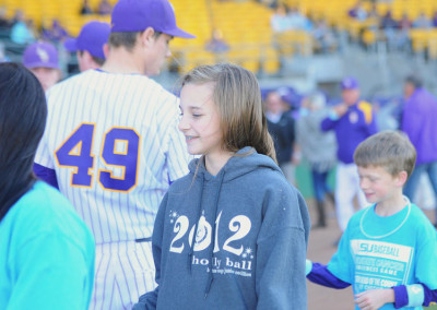 Young girl in a grey hoodie smiling on a baseball field with players and other children in the background.