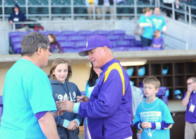 A man in a purple lsu baseball jacket shakes hands with another man in a teal shirt at a sports stadium, with onlookers in the background.