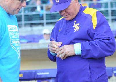 An older man in an lsu baseball uniform signs an autograph at a baseball stadium, interacting with a fan nearby.