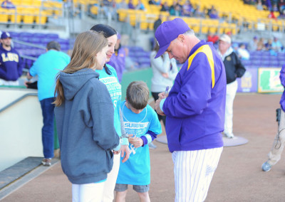 A coach in a purple cap autographs a young boy's shirt at a baseball stadium while a woman smiles at them.