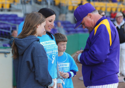 Baseball coach in a purple jacket signing autographs for young fans at a stadium.