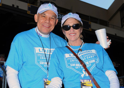 Two adults wearing blue t-shirts and hats with "lsu" logos at a sports event, smiling and holding a drink.