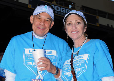 Two adults wearing blue t-shirts promoting prostate cancer awareness at a sporting event, smiling at the camera.