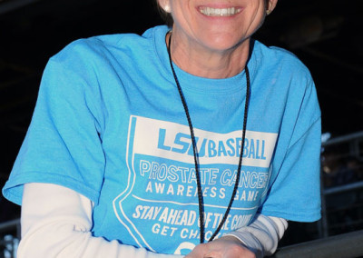 A woman wearing sunglasses, a cap, and a blue t-shirt with text related to prostate cancer awareness, smiling at a sports event.