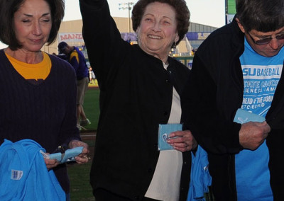 An elderly woman happily waving her hand at a baseball game, flanked by a woman checking her phone and a man holding tickets.