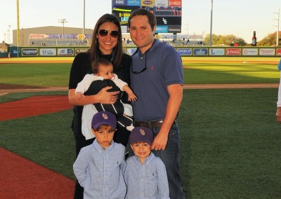 A family of four poses on a baseball field with a scoreboard in the background at alex box stadium.