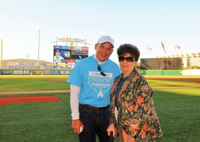 An older couple posing on a baseball field with a scoreboard in the background displaying details of a future game.
