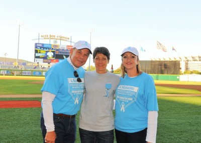 Three adults wearing blue shirts with ribbons stand together on a baseball field, smiling, with stadium seats and advertisements in the background.