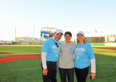 Three people in blue shirts standing on a baseball field, smiling, with a stadium scoreboard in the background.