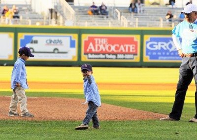 Two young boys in baseball caps and jackets on a baseball field, one gesturing excitedly, with an adult male supervisor nearby.