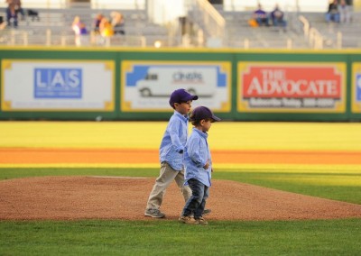 Two boys in baseball caps and jackets standing on a pitcher's mound at a baseball field, with advertising banners in the background.