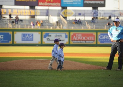 A young child and an adult observing a baseball field with another adult watching, set against advertisements and stadium seating in the background.