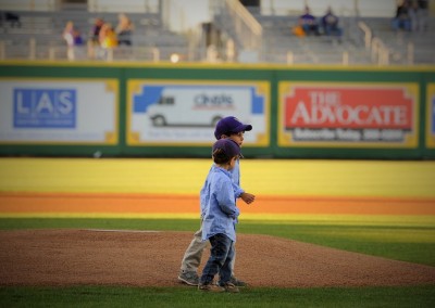 Two young boys in casual clothing stand on a baseball field, one wearing a purple cap, with stadium seats and advertising signs in the background.