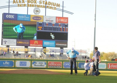 A family walks across a baseball field at alex box stadium, with the scoreboard displaying a game in progress.