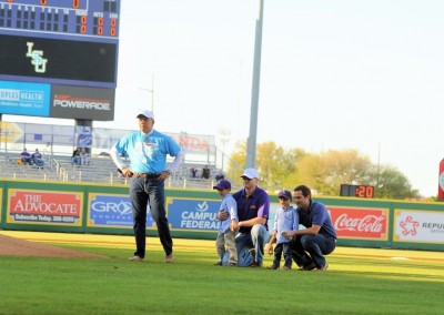 A baseball umpire and several spectators on a field inspecting the ground near first base, with a scoreboard in the background under a clear sky.