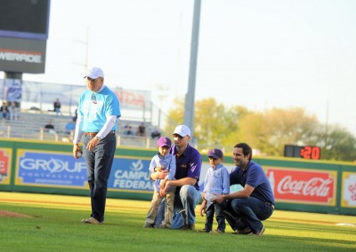 Older man preparing to throw a baseball in a field, accompanied by two younger men and two children, with stadium advertising in the background.