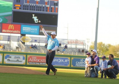 A man throws the first pitch at a baseball game with spectators watching and a scoreboard in the background displaying a 0-0 game.
