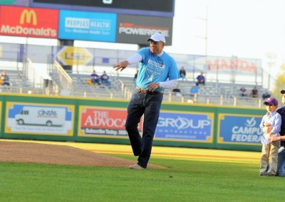 An older man in a blue shirt and jeans throws a ceremonial first pitch at a baseball game, with children watching from the side.