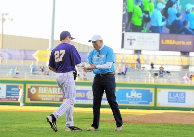 Two men shaking hands on a baseball field, one in a purple jersey and the other in a light blue shirt, with spectators in the background.