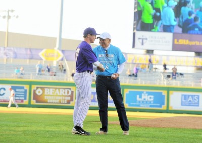 Two men on a baseball field shaking hands, one in a purple lsu jersey and the other in a blue jacket, with spectators in stadium seats in the background.