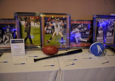 Sports memorabilia auction display with signed photos and a baseball bat on a table, each item accompanied by descriptive tags.