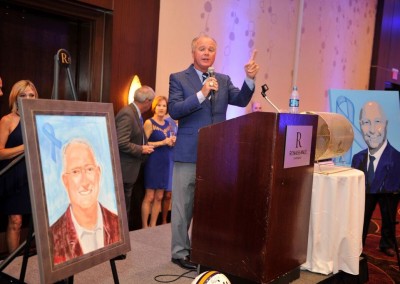 A man speaks at a podium during an event, with portrait paintings displayed beside him.