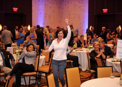 Woman in white shirt cheers joyfully at a corporate event while others chat and drink at tables around her.