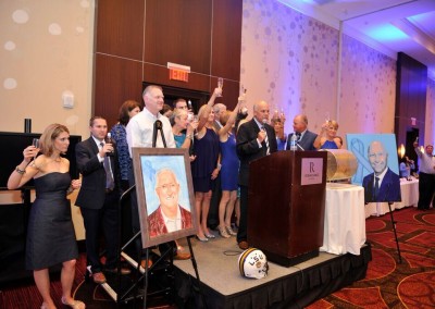 A group of people celebrating at a formal event, with two people speaking at a podium and portraits displayed nearby.