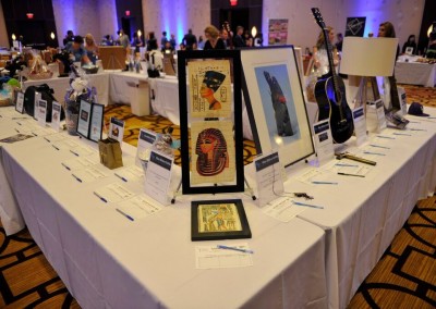 Auction items on display at a charity event, including framed artwork and a guitar, with bid sheets in front of each item.