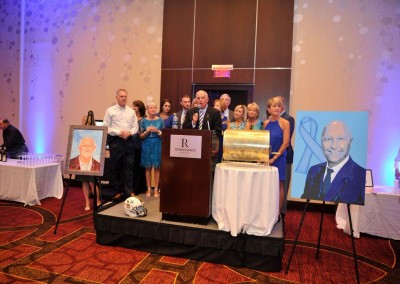 A group of people at a formal event with portraits, a podium, and a cake resembling books on a table.