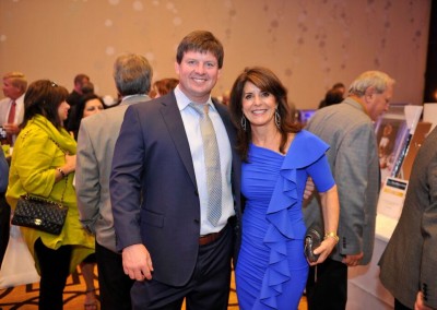 A man and a woman smiling at a formal event, the woman in a blue ruffled dress and the man in a suit.