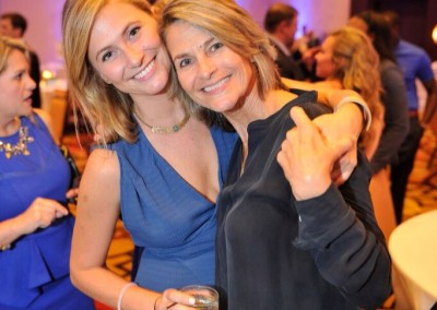 Two women smiling at a social event, one in a blue dress holding a drink, and the other in a black jacket giving a thumbs up.