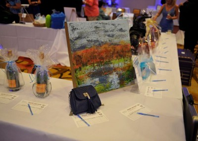 A colorful painting displayed on a table at a charity auction event, flanked by wrapped gift baskets and a small blue purse, with people in the background.