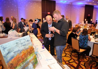 Two men examine items on a table at a charity auction event, with attendees and auction items in the background.