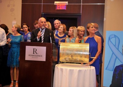 A man speaks at a podium during a charity event, with people in blue attire behind him and raffle tickets visible in a glass bowl.
