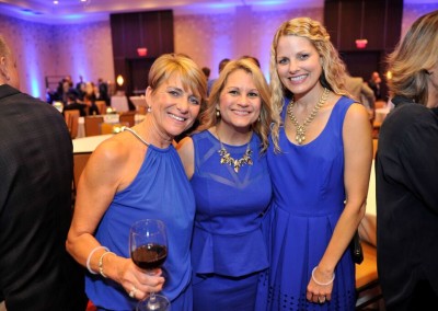 Three women in matching blue dresses smiling at a formal event, one holding a glass of wine.