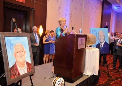 Woman speaking at podium during an event, with portraits on display, other guests looking on, and a decorated venue.