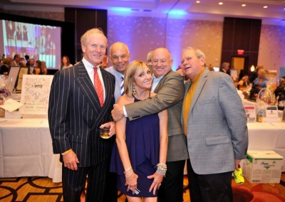 Group of five smiling adults posing at a formal charity event with a crowded room and auction items in the background.