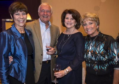 Four smiling elderly adults, two men and two women, posing together at a social event with wine glasses in hand.