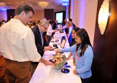 A man interacts with a woman at a buffet table during a conference, with other attendees and a screen in the background.