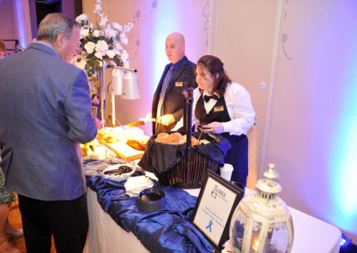A server slices bread for a guest at a banquet, with another server and attendees nearby, under blue-tinted lighting.