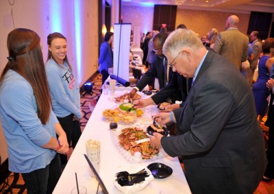 Guests at a networking event serve themselves food from a buffet table while conversing in a hotel conference room.
