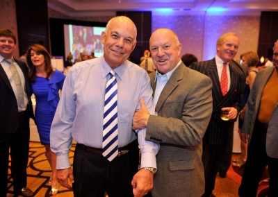 Two smiling older men in business attire with one man's hand on the other's shoulder at a social event with other guests in the background.