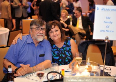 A middle-aged couple smiling at a table during an event, with a sign saying "reserved for the family of joel nasca.