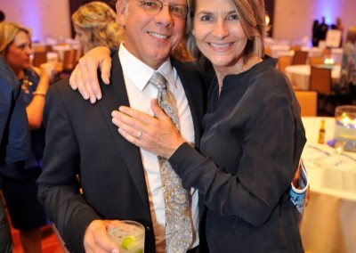 A smiling middle-aged couple posing together at an event, with the man holding a drink.