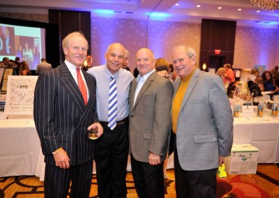 Four smiling men posing together at a formal event with people in the background and items for auction visible on tables.